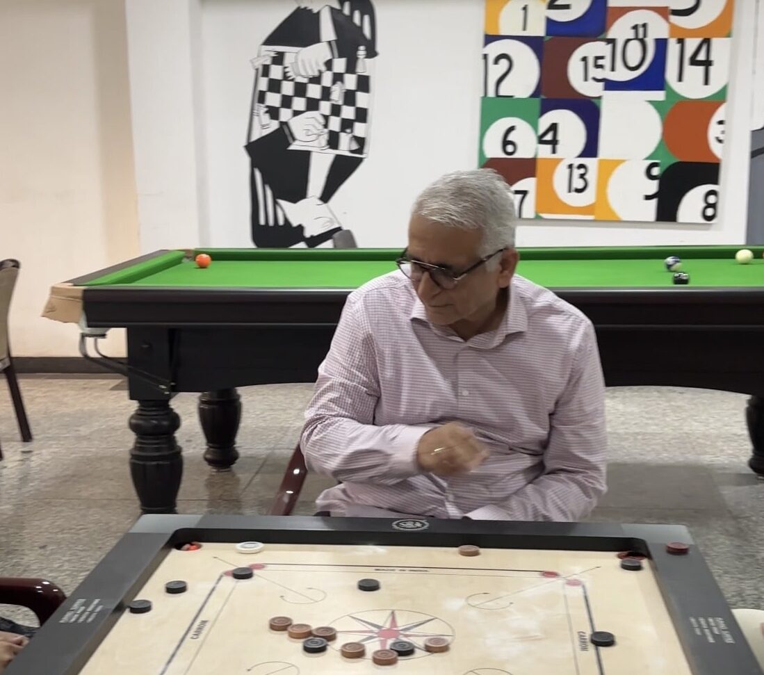 An older adult playing a game of carrom indoors, focusing on the board while sitting near a billiards table