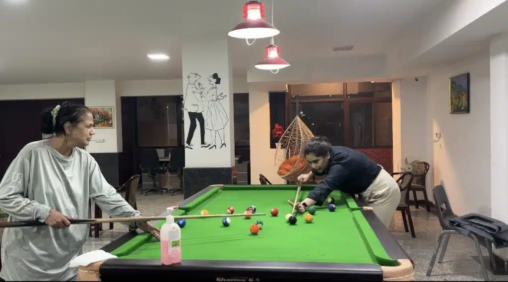 Seniors playing game of pool on a green billiards table inside a well it indoor recreation area.