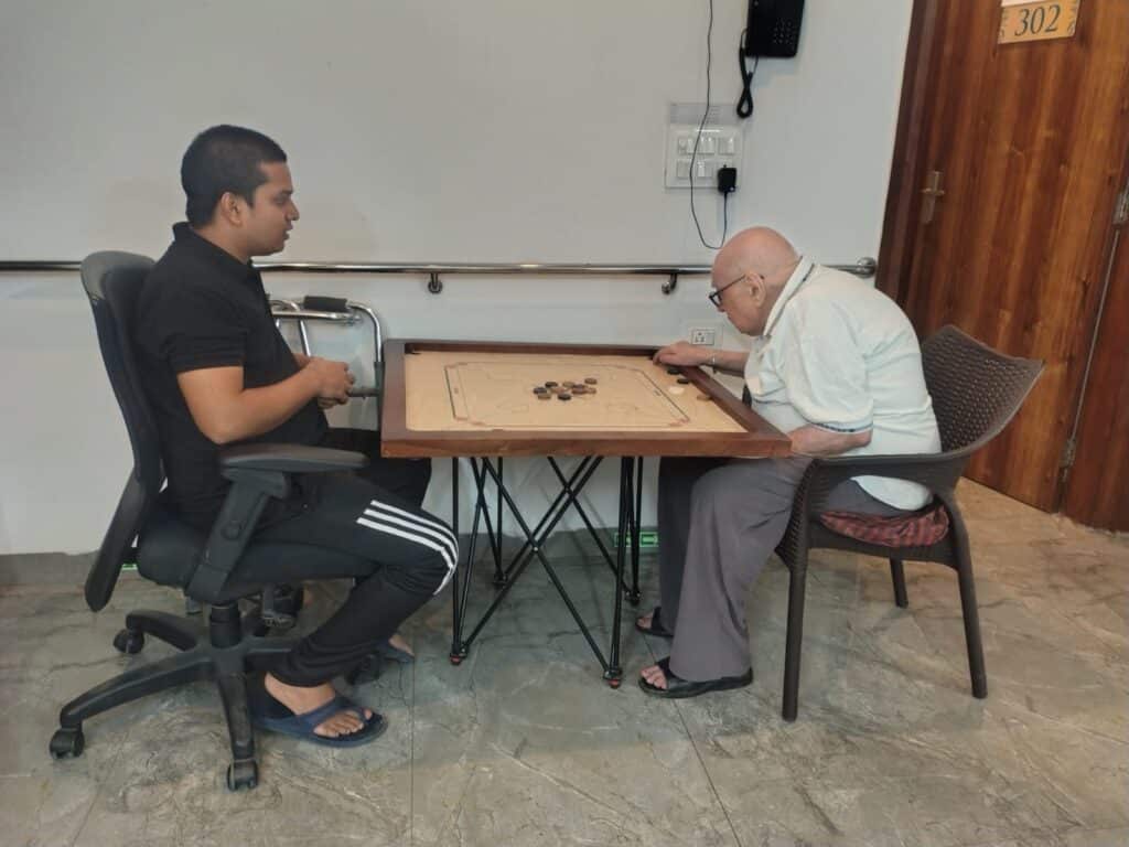 Seniors playing carrom game with a companion at a small indoor table, enjoying a relaxed recreational activity