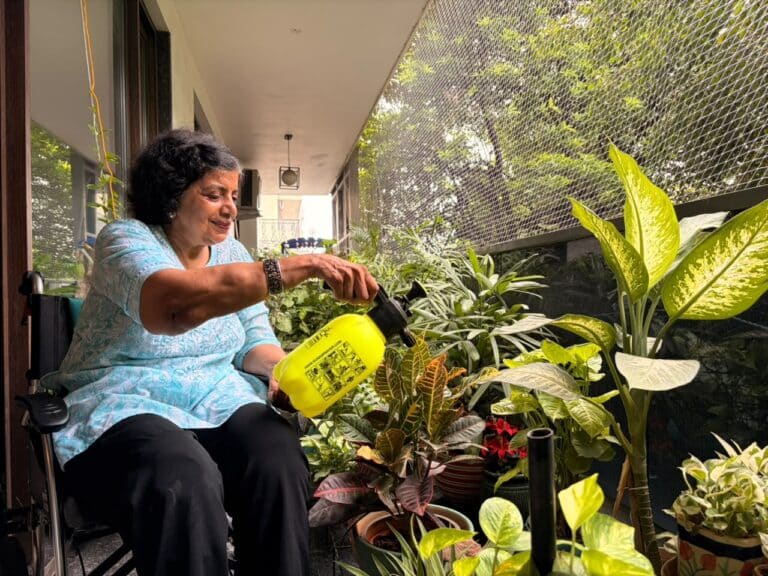 Elderly woman in wheelchair watering potted plants on a green, plant filled balcony at Gracias Living